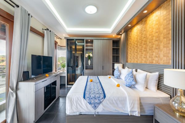 Spacious Balinese-style bedroom with king bed, blue runner, tropical flowers, wooden paneling, TV, and garden view.